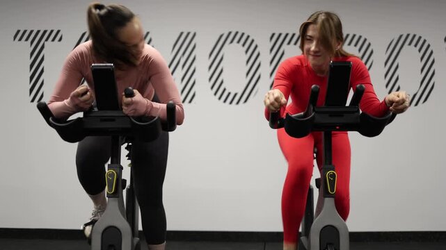Two women compete on stationary bikes pushing their limits and increasing speed while turning a cardio workout into a friendly fitness challenge in the gym