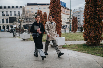 Two business colleagues walk and talk while carrying laptops and documents. The professional pair strolls through a city plaza engaged in conversation. © qunica.com