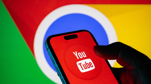 Closeup of a hand holding a smartphone displaying the youtube logo with the google chrome browser icon in the background