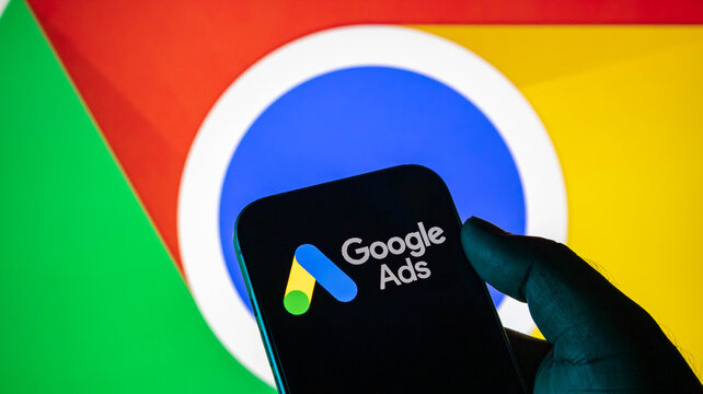 Closeup of a hand holding a smartphone displaying the google ads logo in front of a google chrome browser icon