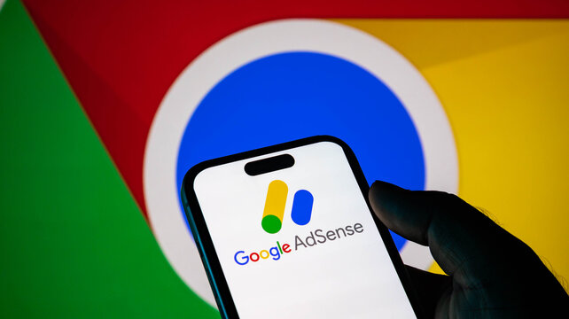 Closeup of a hand holding a smartphone displaying the google adsense logo with the google chrome browser logo in the background