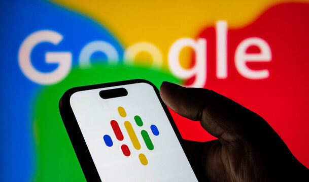 Closeup of a hand holding a smartphone displaying the google podcasts logo in front of a blurred google logo background