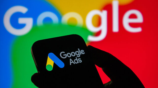 Closeup of a hand holding a smartphone displaying the google ads logo with the google logo in the background