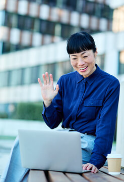 Portrait of a young asian japanese businesswoman woman using a laptop computer having a video call conference and sitting on a bench in a coty park, surrounded by modern corporate office buildings