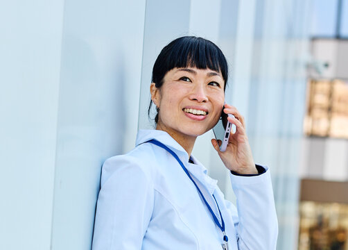 Portrait of an asian female woman doctor or nurse and  health care worker using a smartphone mobile phone holding a cup of coffee   in front of hospital or clinic outside