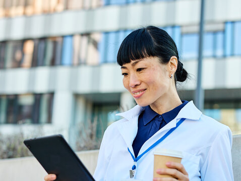 Portrait of an asian female woman doctor or nurse and  health care worker holding a cup of coffee and using a tablet computer in front of hospital or clinic outside