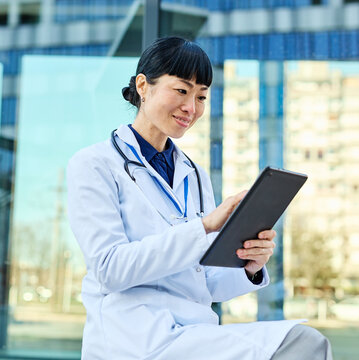 Portrait of an asian female woman doctor or nurse and  health care worker holding a cup of coffee and using a tablet computer in front of hospital or clinic outside