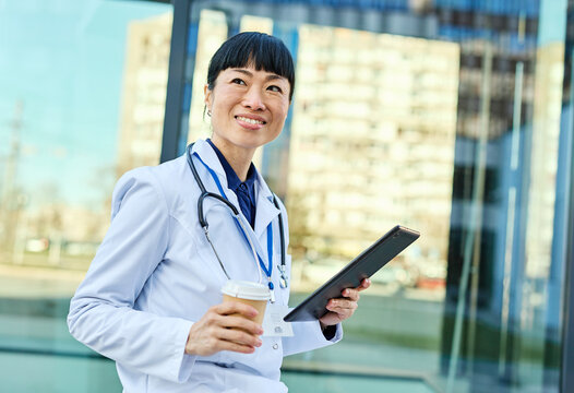 Portrait of an asian female woman doctor or nurse and  health care worker holding a cup of coffee and using a tablet computer in front of hospital or clinic outside