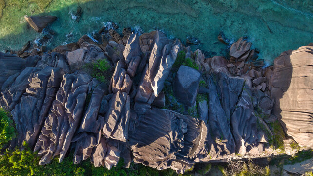 Breathtaking top-down aerial shot of dramatic weathered granite boulders meeting crystal-clear turquoise waters in Anse Grosse Roche, La Digue, Seychelles.