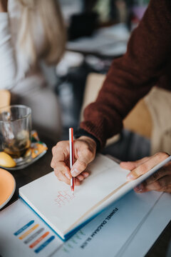 Two business partners jot notes in a notebook while reviewing documents and charts. The hands continue planning and discussing ideas over coffee in a relaxed meeting.