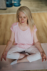 Obraz premium Mature woman with closed eyes practices seated meditation on a yoga mat. She sits cross-legged in a calm home setting focusing on breath and relaxation.
