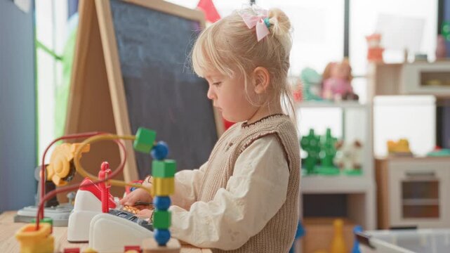 Little blonde girl playing with a bead maze and toy cash register, hand touching blocks in a building; quiet concentration.