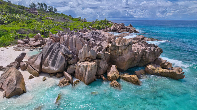 High-angle drone shot of dramatic granite boulders, white sandy beach, and vibrant tropical forest meeting the turquoise ocean at Anse Cocos, La Digue, Seychelles. Beautiful nature background