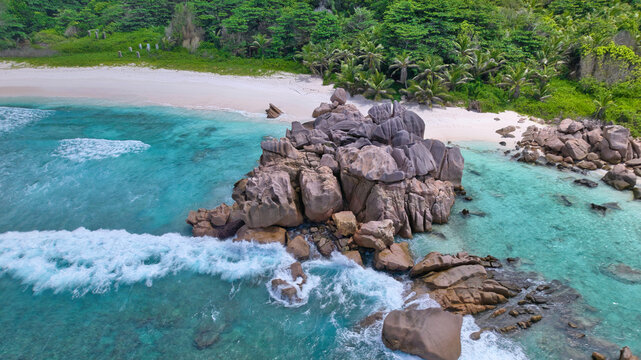 High-angle drone shot of dramatic granite boulders, white sandy beach, and vibrant tropical forest meeting the turquoise ocean at Anse Cocos, La Digue, Seychelles. Beautiful nature background