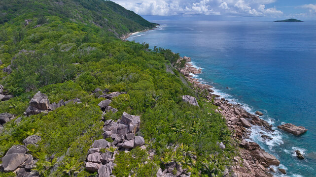 Stunning high-angle drone shot capturing the wild, forested coastline and granite boulders meeting the deep blue ocean waves near Anse Cocos, La Digue, Seychelles.