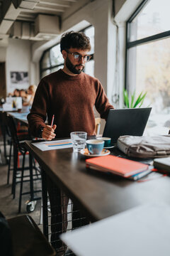 A man works on a laptop at a cafe table while reviewing printed charts and taking notes. The casual business scene shows remote work and focused productivity in a bright coworking-style space.