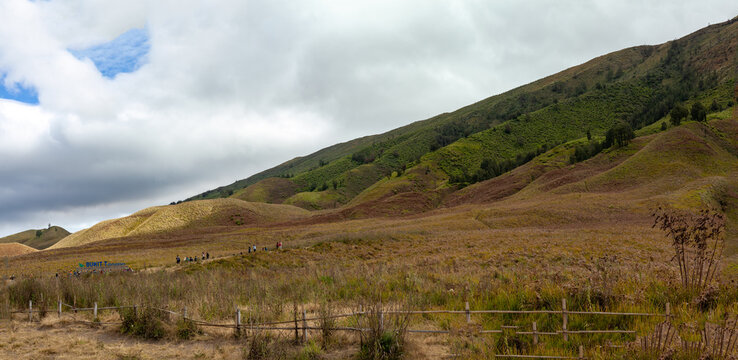 Panoramic view of Bromo Tengger Semeru national park landscape with volcanic hills and green savanna under cloudy sky.