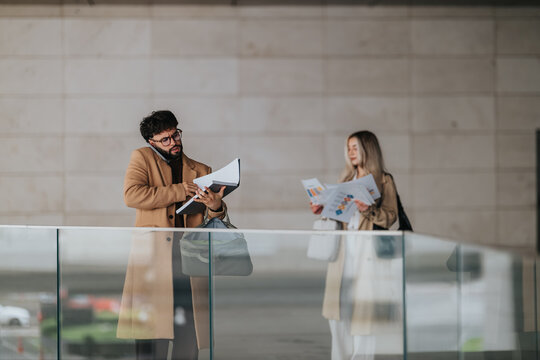 Two business colleagues stand in a modern atrium reviewing documents while one speaks on a phone. The pair examine charts and papers during a brief work discussion in a glass-walled public space.