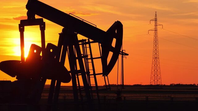 Silhouette of oil pump jack against a colorful beautiful sky with glowing clouds at sunset, high voltage electric pole in background