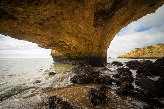 View of a massive limestone rock arch over a sandy beach with waves crashing against dark rocks in Albufeira, Faro District, Portugal.