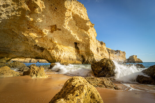 View of golden limestone cliffs and a natural sea cave with waves crashing against rocks on a sandy beach under a clear blue sky in Albufeira, Faro District, Portugal.