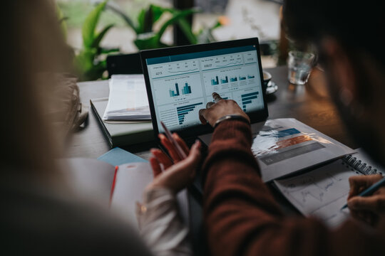 Two business colleagues analyze charts and data on a laptop screen while taking notes. The scene shows a collaborative meeting focused on analytics and planning.