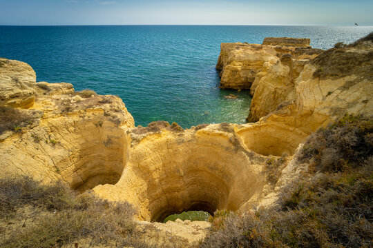 View of the golden limestone cliffs and deep sinkholes overlooking the turquoise Atlantic Ocean under a clear sky in Albufeira, Faro District, Portugal.
