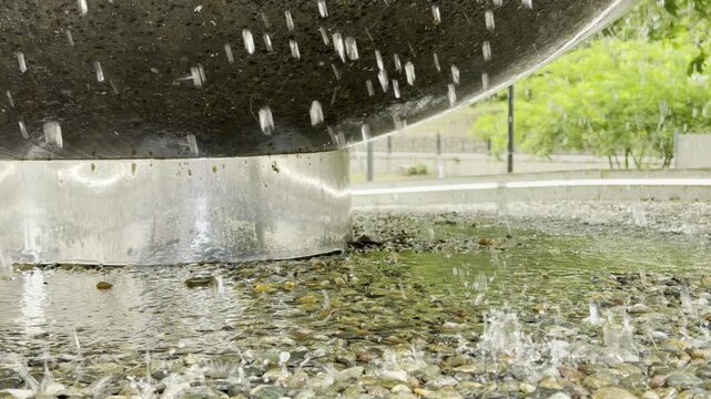 Water drips rapidly from a heavy stone fountain into a wet pebble pool. This lively splashing creates a deeply refreshing and cheerful mood in the green park.