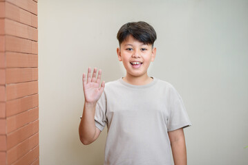 Cheerful Asian preteen student boy waving hand and smiling warmly. Great concept for back to school welcoming, friendly youth communication, and educational advertisements © Kornkanok