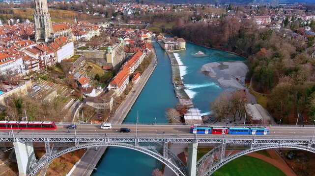 4K aerial drone footage of Bern, Switzerland - Kornhausbr&uuml;cke bridge with red and blue trams crossing the turquoise Aare River, Bern M&uuml;nster Gothic tower, and UNESCO old town in winter.
