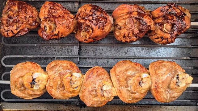 Top-down view inside an oven showing two parallel horizontal skewers: the main subject is a row of five deeply caramelized pork knuckles across