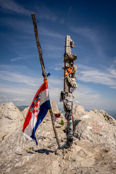 View of the Croatian flag and mementos on a wooden pole at the summit of Tulove Grede with rocky limestone peaks under a blue sky Jasenice, Zadar County, Croatia.