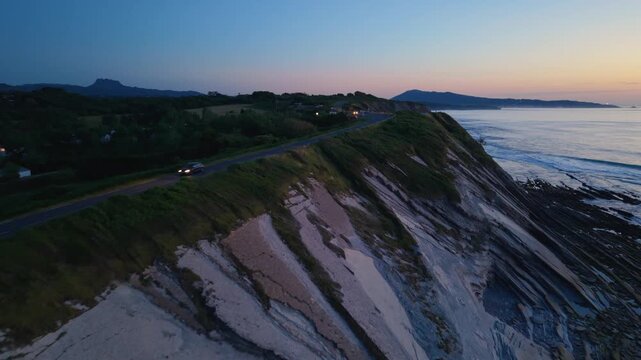 Aerial views show the Basque Country's coastal road at dusk. The cliffs and ocean are illuminated in fading light
