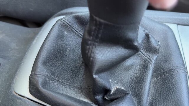 Manual gear stick is shifted inside a passenger car, close-up of textured black leather gear boot and center console, driving action, transmission control.