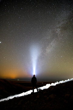 View of a person standing on a rocky ridge at night, pointing a bright beam of light towards the starry sky and Milky Way galaxy El Cotillo, Canary Islands, Spain.