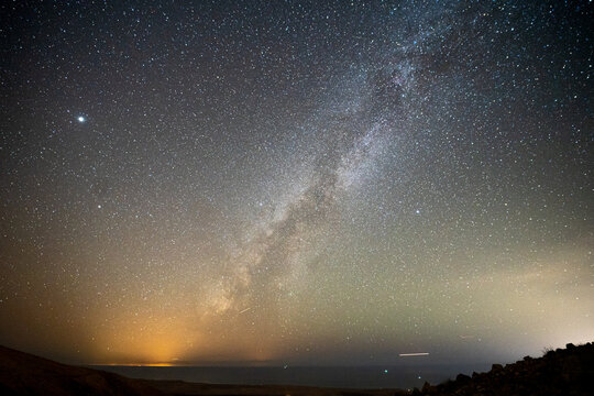 View of the Milky Way galaxy stretching across a starry night sky over a dark horizon with distant lights and rocky terrain in El Cotillo, Canary Islands, Spain.