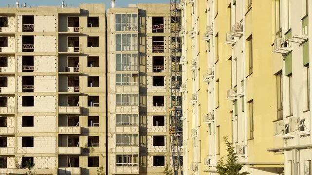 A large concrete apartment building stands incomplete at a busy construction site. It rises next to a finished, pale yellow residential complex equipped with air conditioners.