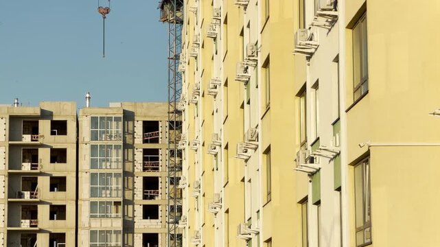 A tall yellow residential building features rows of windows and air conditioners. Next to it, a new concrete structure is under construction. A crane hook hangs down against a clear blue sky.