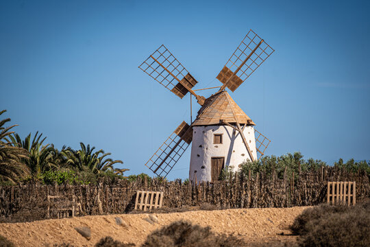 View of a traditional white windmill with wooden sails and a conical roof standing behind a rustic fence under a clear blue sky in El Cotillo, Canary Islands, Spain.