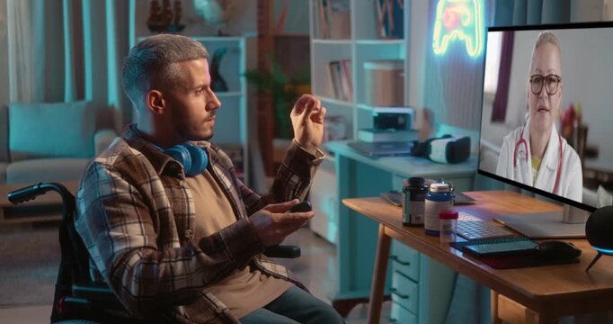 Young Man in wheelchair holds medication while consulting a doctor through a video call on computer at home. Telemedicine and remote healthcare concept.