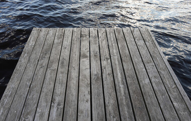 Fototapeta premium A weathered gray wooden dock extends into calm water, highlighting textured planks, gentle ripples and a quiet waterfront mood ideal for relaxation, solitude, boating, fishing and nature scenes