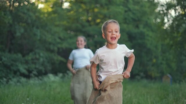 Boy runs ahead in summer competition. Girl jumps in sack race outdoors. Kid plays game with joy. Jumping energy in children. Girl catches up in summer game. Outdoors play kid, boy, child having fun.