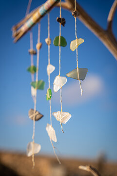 View of a decorative wind chime made of sea glass and stones hanging from a branch under a bright blue sky in El Cotillo, Canary Islands, Spain.