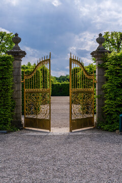Opened golden gates with intricate scrollwork stand between stone pillars, leading to an open gravel path within Herrenhausen Gardens.