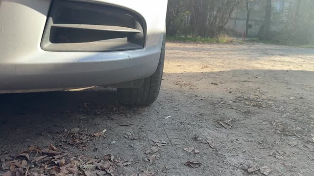 Car wheel moves across a rough gravel surface beneath the front bumper, close-up of rolling tire, suspension movement, road vibration, vehicle travel.