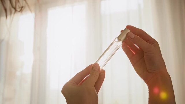 Caucasian woman hands holding and rotating small glass perfume vial against window background. Clip is perfect for cosmetics marketing and beauty product promotions