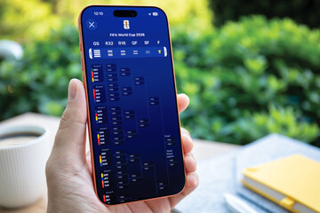 Naklejka premium Man hands holding mobile phone Apple iPhone 17 Pro Max Cosmic Orange and application Apple Sports and FIFA World Cup 2026 on the screen ın the desk background