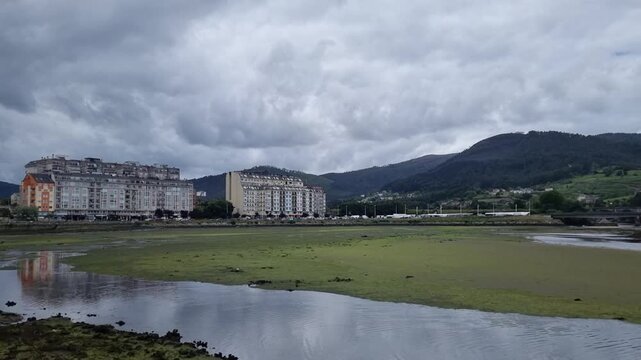 Wide panoramic view of Viveiro, Spain, featuring the estuary, coastal buildings on green hills, and a cloudy blue sky.
