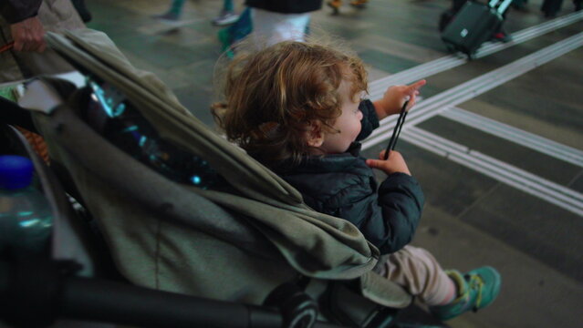 Toddler seated in stroller at busy station looking upward with curiosity as travelers pass by evoking wonder transition public travel movement and early social awareness
