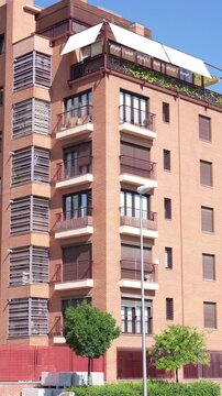Vertical view of a modern residential apartment building featuring balconies, terraces, awnings and brick facade. Urban housing architecture with residential details on a sunny day in Madrid, Spain.